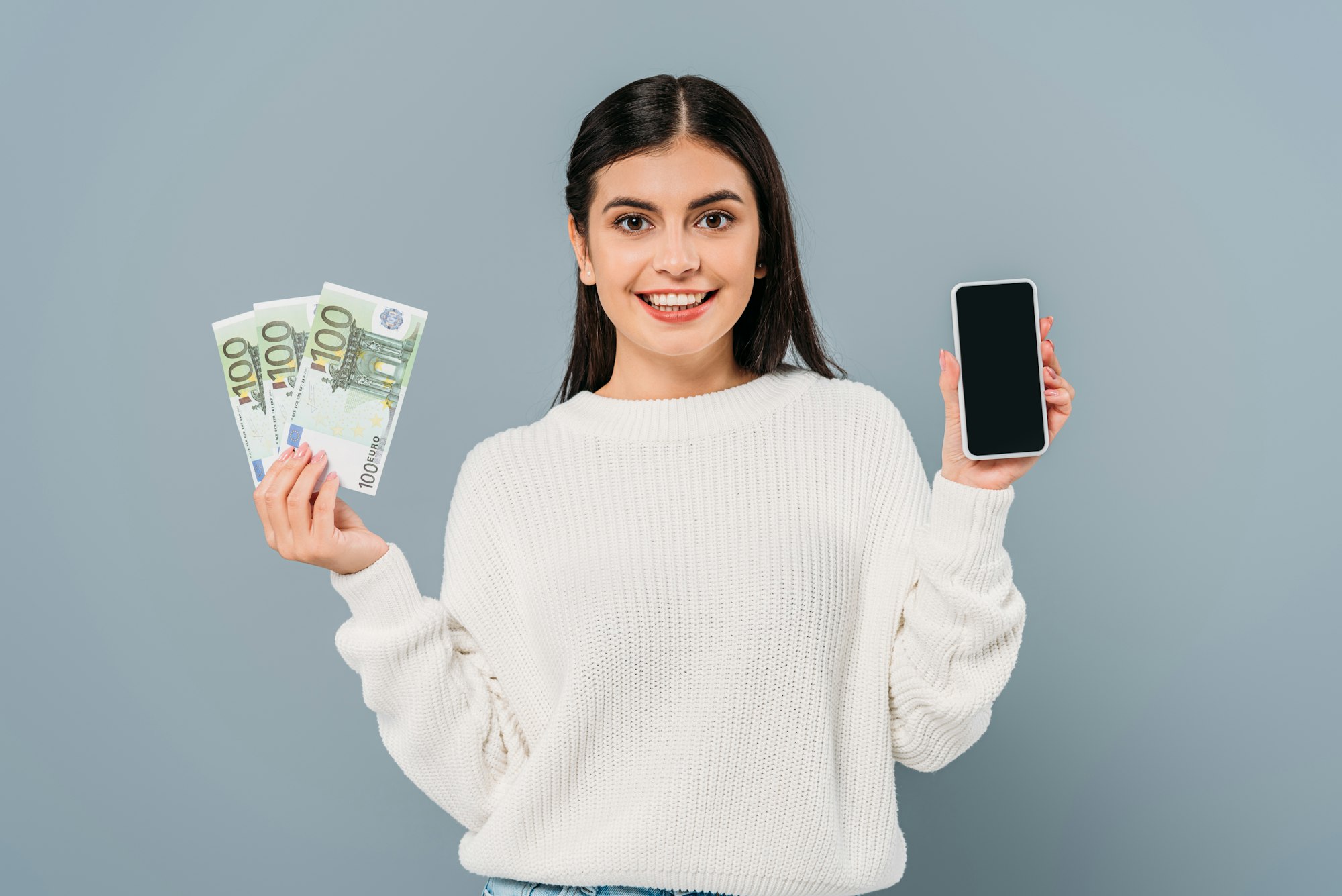 smiling pretty girl in white sweater holding euro banknotes and smartphone with blank screen